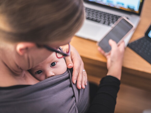 Une mère regarde son bébé en écharpe, un téléphone à la main et un ordinateur portable sur le bureau. 