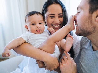 La maman tient le bébé dans ses bras en souriant. Le bébé a le pied sur le nez de son père.