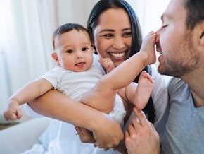 Die Mutter hält das Baby und lächelt. Das Baby hat einen Fuß auf der Nase des Vaters.