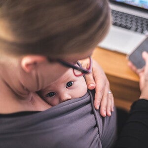 Mutter mit Handy in der Hand und Laptop auf dem Tisch sieht ihr Baby in der Trage an