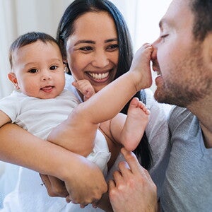 Die Mutter hält das Baby und lächelt. Das Baby hat einen Fuß auf der Nase des Vaters.